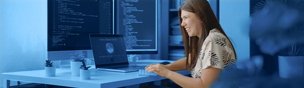 A smiling woman coding on multiple computer screens with a laptop and plants on her desk.